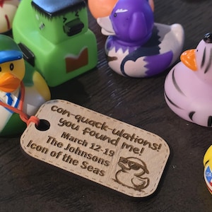Puede incluir: Una etiqueta de madera con el texto "con-quack-ulations! you found me! March 12-19 The Johnsons Icon of the Seas" y una imagen de dibujos animados de un pato de goma con gafas de sol. La etiqueta est&aacute; unida a un pato de goma verde con una gorra de b&eacute;isbol azul.