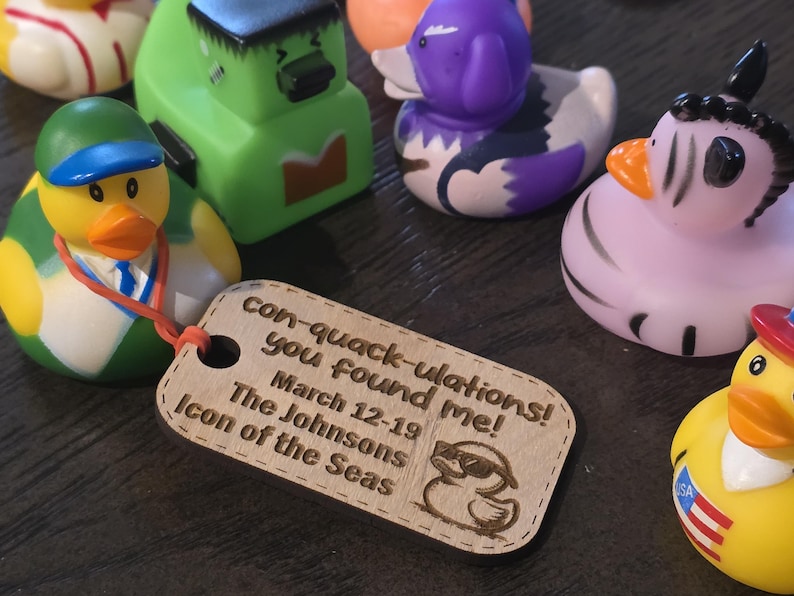 Puede incluir: Una etiqueta de madera con el texto "con-quack-ulations! you found me! March 12-19 The Johnsons Icon of the Seas" y una imagen de dibujos animados de un pato de goma con gafas de sol. La etiqueta est&aacute; unida a un pato de goma verde con una gorra de b&eacute;isbol azul.