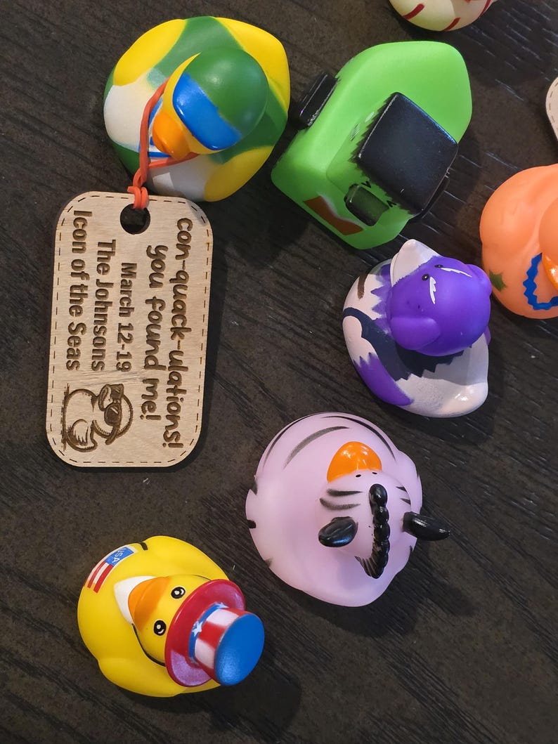 Puede incluir: Un letrero de madera con el texto "con-quack-ulations! you found me! March 12-19 The Johnsons Icon of the Seas" y un pato de dibujos animados con gafas de sol. Hay varios juguetes de patos de goma en el fondo, incluyendo un barco verde, un pato morado con un patr&oacute;n de cebra, un pato amarillo con un sombrero azul y blanco, y un pato amarillo con un patr&oacute;n verde y blanco.