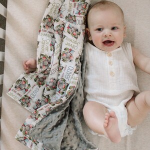 May include: A baby boy is lying on a white and gray blanket with a colorful pattern. The blanket has the names "Oliver" and "James" repeated throughout the pattern. The baby is wearing a white sleeveless onesie with buttons down the front.