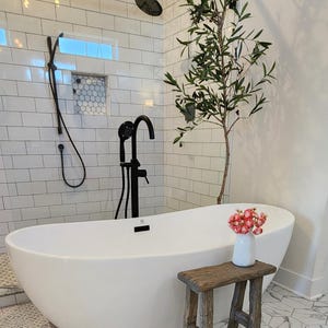May include: A modern bathroom with a white, oval-shaped bathtub. The bathroom features a black showerhead and faucet, white subway tile walls, and a small wooden stool. A vase of pink flowers sits on the stool.
