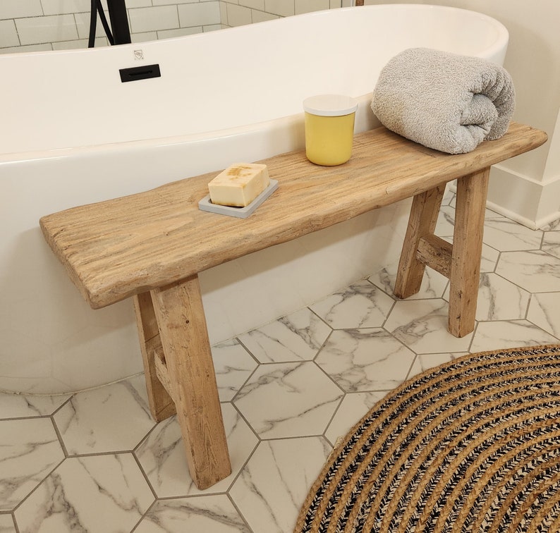 May include: A rustic wooden bench with a natural finish, holding a gray towel, a yellow container, and a bar of soap on a gray soap dish. The bench is positioned near a white bathtub.