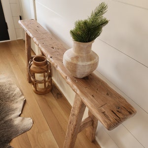 May include: A rustic wooden bench with a distressed finish. A beige vase with green foliage sits on the bench. A wooden lantern with a rope handle and candles is also on the bench. The bench is placed against a white wall.