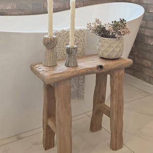 May include: A rustic wooden stool with a natural finish, holding two cream-colored candles in decorative holders. A small vase with dried flowers sits on the stool. The background features a white bathtub and brick wall.