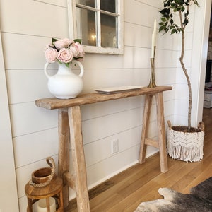 May include: A rustic wooden console table with a distressed finish. A white vase with pink roses sits on the table, along with a white rectangular tray. A wooden lantern and a macrame basket with a plant are also visible.