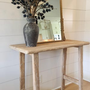 May include: A rustic wooden console table with a distressed finish, topped with a gray vase holding dark blue decorative branches. A gold-framed mirror leans against the wall behind the table.