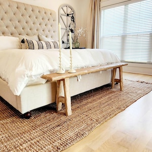 May include: A bedroom scene featuring a tufted headboard, white bedding, and a wooden bench at the foot of the bed. A woven jute rug and two tall candles add to the room's cozy, neutral aesthetic. The room has a window with blinds.