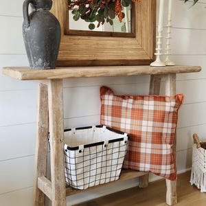 May include: A rustic wooden console table with a distressed finish. It features a decorative mirror, candles, and a vase with branches. A plaid pillow and a wire basket add to the farmhouse aesthetic. The table is 48 inches long.