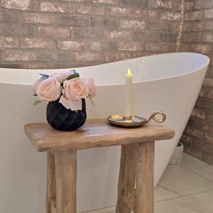 May include: A rustic wooden stool holds a black geometric vase filled with pink roses, a gold candle holder with a lit candle. A white bathtub and brick wall are in the background.