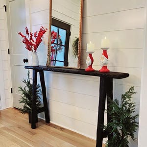 May include: A rustic black console table with a gold-framed mirror, white candles, and red accents. The table is flanked by small evergreen trees in pots. The wall is white with horizontal wood paneling, and the floor is light-colored wood.