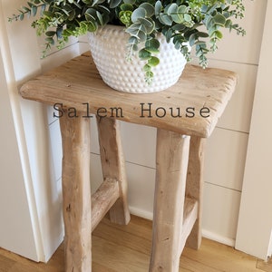 May include: A rustic wooden stool with a square top and four legs supports a white, textured ceramic pot filled with green and gray eucalyptus. The text "Salem House" is visible on the stool.