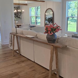 May include: A rustic wooden console table with a natural finish stands in front of a white sofa. A vase of pink flowers sits on the table. The room has hardwood floors and large windows.