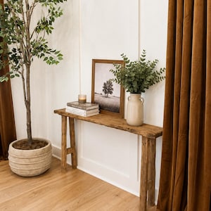May include: A rustic wooden console table with a natural finish, featuring a rectangular top and four straight legs. On the table sits a stack of two books, a small glass candle, a framed black and white landscape print, and a beige ceramic vase.