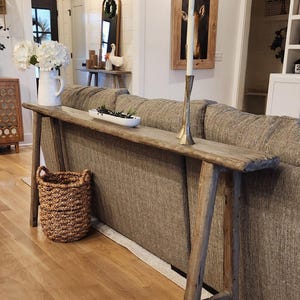 May include: A rustic wooden console table with a natural finish, placed behind a gray sofa. The table has a long, narrow top and angled legs. A woven basket and decorative items sit on the table. The room has hardwood floors and white walls.