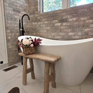 May include: A rustic wooden bench with four legs sits in front of a white bathtub. A woven basket with pink and burgundy flowers rests on the bench. The bathroom has a brick wall and a large window.