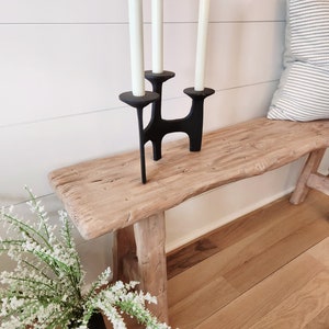 May include: A black metal candle holder with three white candles on a rustic wooden bench. The candle holder has a unique, modern design. A vase of white flowers sits to the left of the bench. The background is a white wall with horizontal wood paneling.