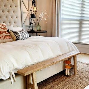 May include: A bedroom scene featuring a tufted headboard, a wooden bench, and a jute rug. The bed is covered with white bedding and decorative pillows. A small table holds a lamp and vase. Decorative mushrooms are placed near the bench.