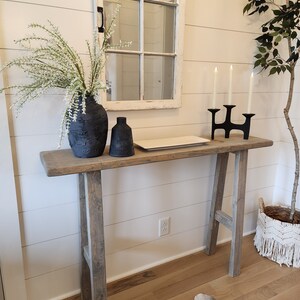 May include: A rustic wooden console table with a distressed finish. It features a black vase with white floral stems, a small black bottle, a white rectangular tray, and a black candle holder with three lit candles. A decorative mirror hangs above.