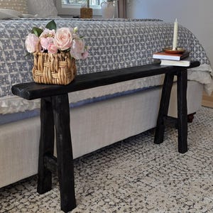 May include: A black wooden bench with a rustic design, featuring a rectangular top and A-frame legs. A woven basket with pink roses sits on the bench, along with books and a candle. The bench is placed in front of a bed.