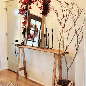 May include: A rustic wooden console table with a large rectangular mirror adorned with red leaves. A black candelabra with white candles sits on the table. A decorative skeleton sits on the table. A woven basket holds branches.