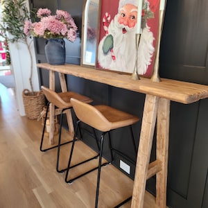 May include: A rustic wooden console table with two leather-topped bar stools. The table is decorated with a Santa Claus painting, brass candlesticks, and a mirror. The stools have black metal legs. The floor is light-colored wood.
