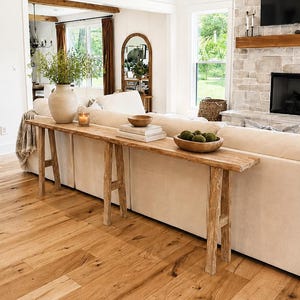 May include: A long, narrow, light-toned wooden console table with a rustic, weathered texture and straight legs, positioned behind a beige sofa. The table surface holds a large ceramic vase with greenery, a candle, books, and wooden bowls.