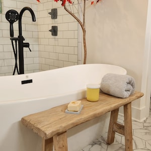 May include: A rustic wooden bench with a rolled gray towel, a yellow jar, and a bar of soap on a gray dish. A white bathtub and black shower fixtures are in the background. A decorative tree with red leaves adds a pop of color.
