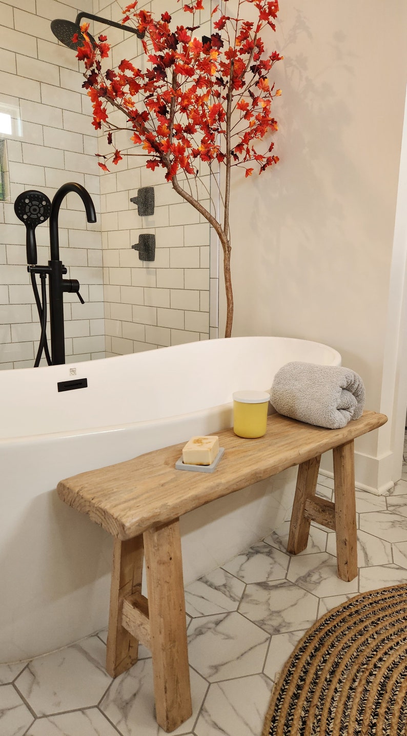 May include: A rustic wooden bench with a rolled gray towel, a yellow jar, and a bar of soap on a gray dish. A white bathtub and black shower fixtures are in the background. A decorative tree with red leaves adds a pop of color.