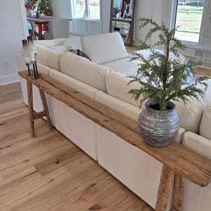 May include: A rustic wooden console table with a live edge design. The table is placed behind a white sofa and has a decorative plant in a silver pot. The room has hardwood floors and large windows.