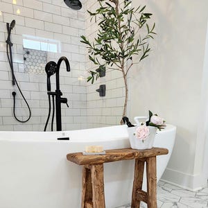 May include: A rustic wooden bench sits in front of a white bathtub. The bathroom features a black shower fixture and a small tree. The floor is tiled with a white and gray hexagonal pattern. A small white bucket with pink flowers is next to the tub.