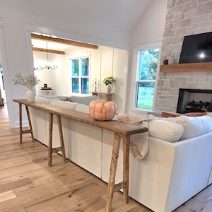 May include: A long, rustic wooden console table with a natural finish. A light orange pumpkin sits on the table, which is placed behind a white sofa. The room has hardwood floors and a stone fireplace.