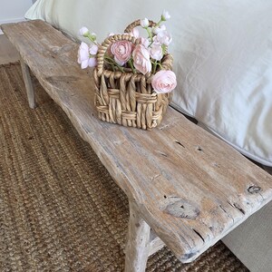 May include: A rustic wooden bench with a woven basket filled with pink flowers. The bench is placed on a woven jute rug. The scene is set in a bedroom with a white bedspread.