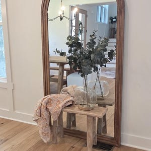 May include: A tall, arched floor mirror with a medium-toned wooden frame. In front of the mirror sits a small, rustic wooden bench holding a clear glass vase filled with green eucalyptus branches and a folded, patterned peach-colored textile.