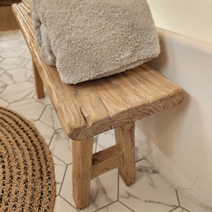 May include: A rustic wooden bench with a light gray towel folded on top. The bench has a natural wood finish and is supported by four sturdy legs. The towel appears soft and plush. The bench is placed near a white bathtub.