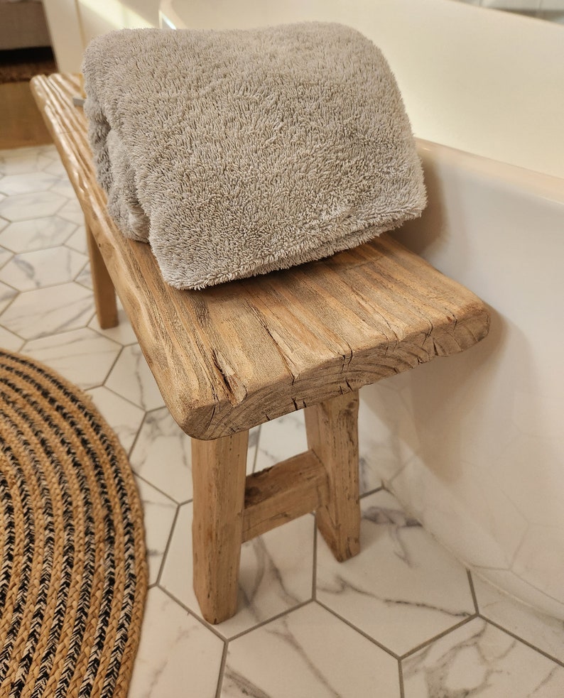 May include: A rustic wooden bench with a light gray towel folded on top. The bench has a natural wood finish and is supported by four sturdy legs. The towel appears soft and plush. The bench is placed near a white bathtub.