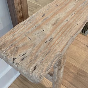 May include: A long, narrow wooden bench crafted from light-toned, weathered timber with a visible grain, natural knots, and a rough, textured surface. The bench features four sturdy, cylindrical legs and a thick, rectangular seat with rounded edges.