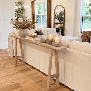 May include: A long, narrow wooden console table with four angled legs positioned behind a white sofa. The table surface holds a textured gray vase with dried foliage, a small wooden bowl with white pumpkins, and two glass candle holders.