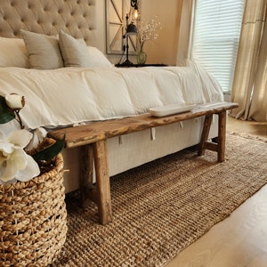 May include: A rustic wooden bench with a natural edge sits at the foot of a bed with a tufted headboard. A woven jute rug and a basket of flowers add to the room's cozy, neutral aesthetic. The room is bathed in natural light.