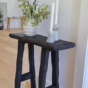 May include: A black wooden console table with a rectangular top and four sturdy legs. The table is adorned with a white vase holding green foliage, and two white candles in decorative holders. The table's dimensions are approximately 48 inches long, 12 inches wide, and 30 inches high.