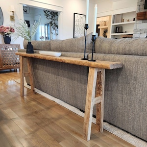 May include: A rustic wooden console table with a natural edge, supported by A-frame legs. The table is placed against a gray fabric sofa. Two black candle holders with white candles are on the table. A vase of white flowers and a decorative bowl are also on the table.