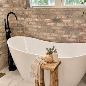 May include: A rustic wooden stool stands in front of a white freestanding bathtub. On the stool sits a beige textured towel, a small candle in a glass jar, a natural sea sponge, a small ceramic vase with greenery, and a beige soap dispenser.
