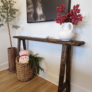 May include: A rustic, dark-stained wooden console table with a live edge design. A black and white horse portrait hangs above the table. A white vase with red berries and a decorative basket with a plush toy are on the table.