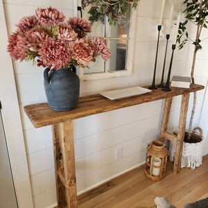 May include: A rustic wooden console table with a distressed finish. It features a dark gray vase filled with pink flowers, black candle holders, and a decorative white tray. A wooden lantern and a macrame basket add to the farmhouse aesthetic.
