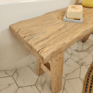 May include: A rustic wooden bench with a natural finish, placed near a white bathtub. A bar of soap rests on a small gray tray on the bench. The bench has a weathered appearance, adding a touch of natural decor to the bathroom.