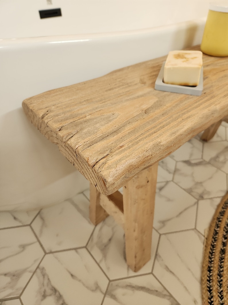 May include: A rustic wooden bench with a natural finish, placed near a white bathtub. A bar of soap rests on a small gray tray on the bench. The bench has a weathered appearance, adding a touch of natural decor to the bathroom.