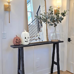 May include: A Halloween-themed entryway featuring a black console table with two decorative pumpkins and a gold-framed mirror. The table is adorned with a clear glass vase holding greenery, and the walls are decorated with sconces.