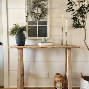 May include: A rustic wooden console table with a distressed finish. A black textured vase holds greenery. A window frame with a wreath hangs on the white shiplap wall. Two gold candlesticks and a wooden lantern complete the decor.