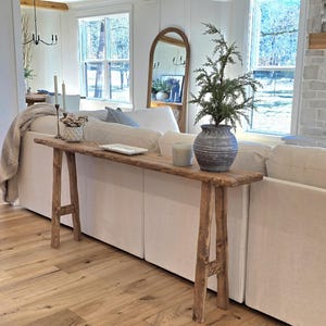 May include: A rustic, wooden console table with a long, narrow top and angled legs. The table is placed behind a white sofa and decorated with a vase, candles, and a small plant. The room has hardwood floors and large windows.