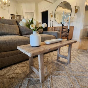 May include: A rustic wooden coffee table with a distressed finish, holding a white vase of white tulips and a white rectangular dish. The table sits on a patterned beige rug in a living room setting.