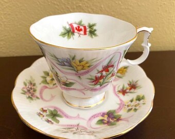 May include: A white teacup and saucer set with a gold rim. The teacup features a Canadian flag and floral designs. The saucer has a matching floral pattern with pink ribbons and gold trim. A decorative tea set.